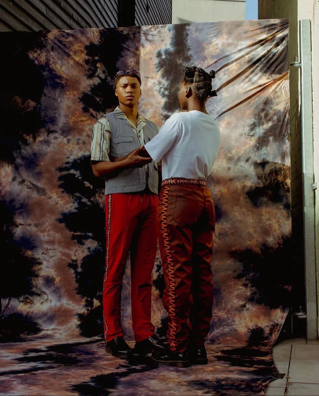 Two young people with separte hairstyles stand in front of a printed backdrop of a cloudy sky. They are facing and holding each other's hands, with one person looking directly at the camera.