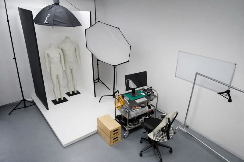 Photography studio with two mannequin models, umbrella and octagonal softbox, white backdrop, computer and photography equipment on a rolling cart, chair, wooden boxes, and studio lighting and support equipment.