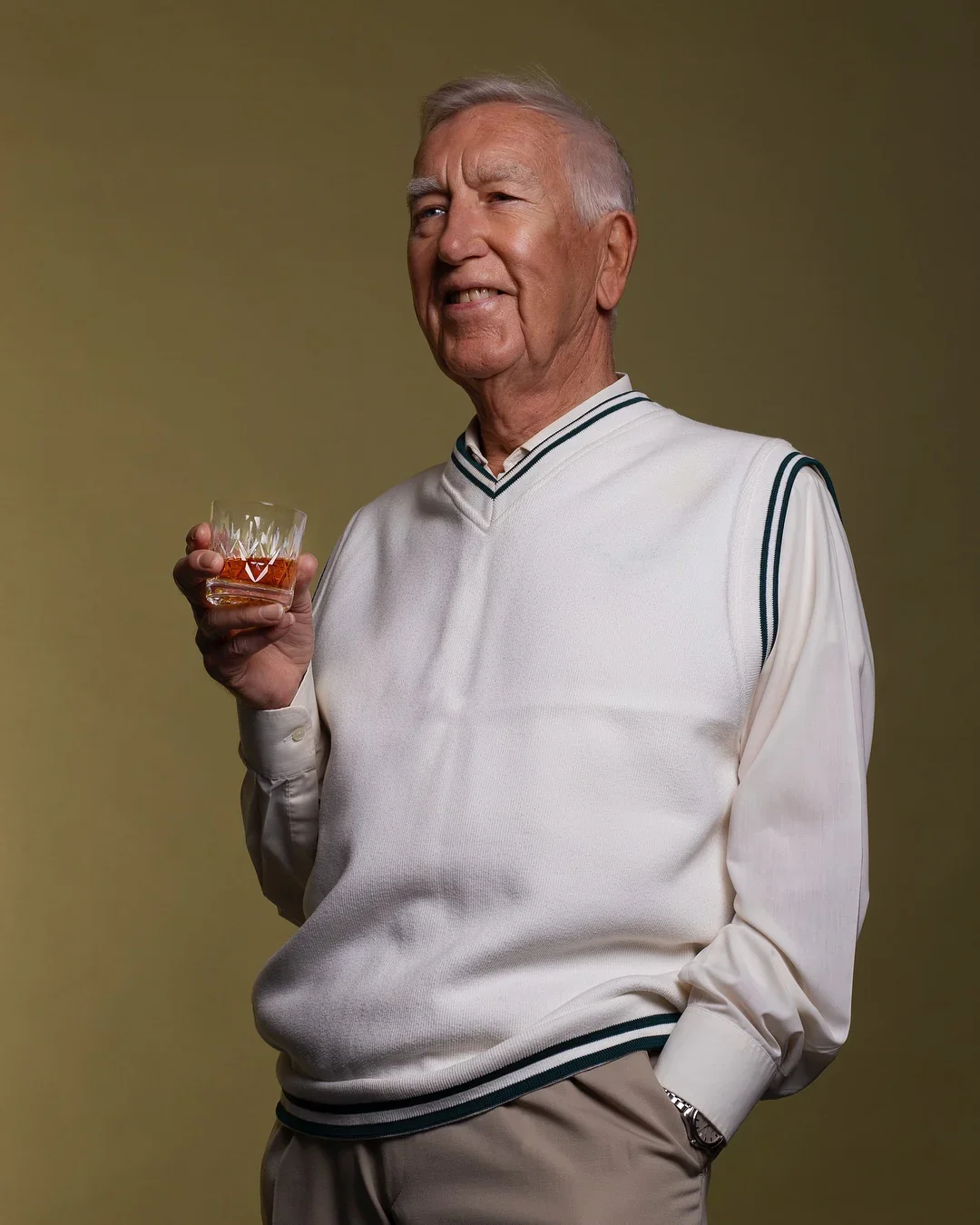 Older man with gray hair smiling while holding a glass of whiskey, dressed in a white sweater vest with green and black stripes over a white shirt, standing against a neutral background.