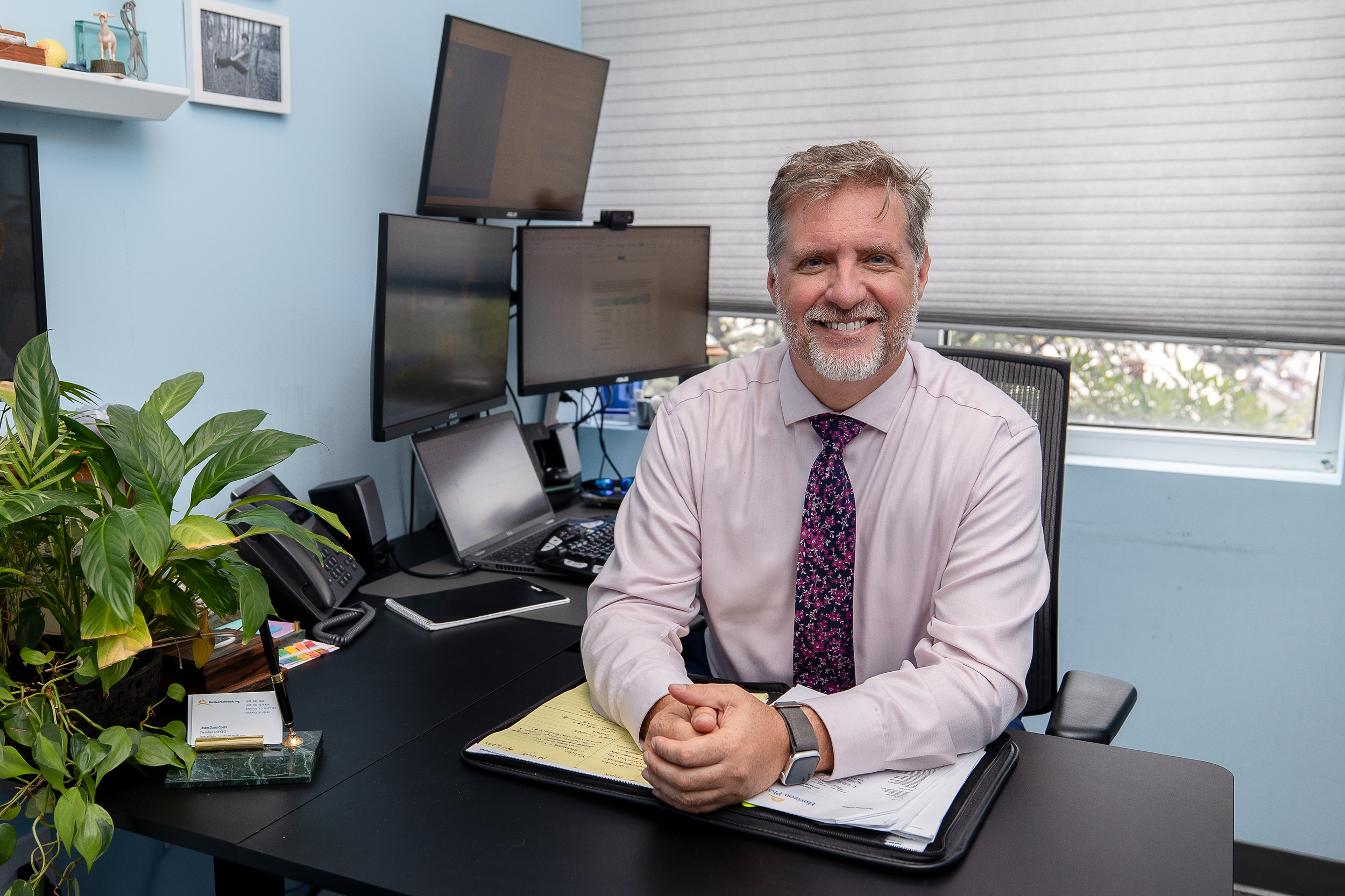 Smiling man sitting at a desk in an office, surrounded by multiple computer monitors, a laptop, office phone, and a green plant.