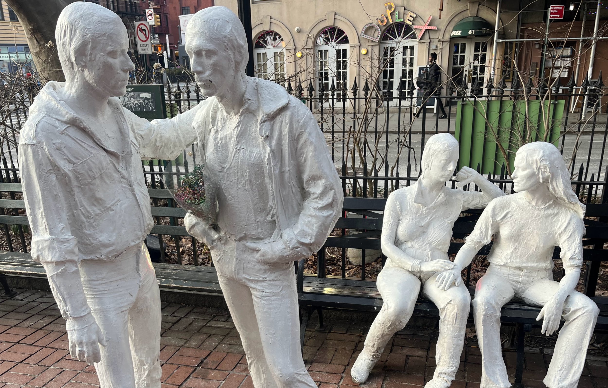 Four white painted statues of young people on a city NYC; two standing and two sitting on a bench, with buildings and signs in the background. Gay Liberation Monument by George Segal in Christopher Park.