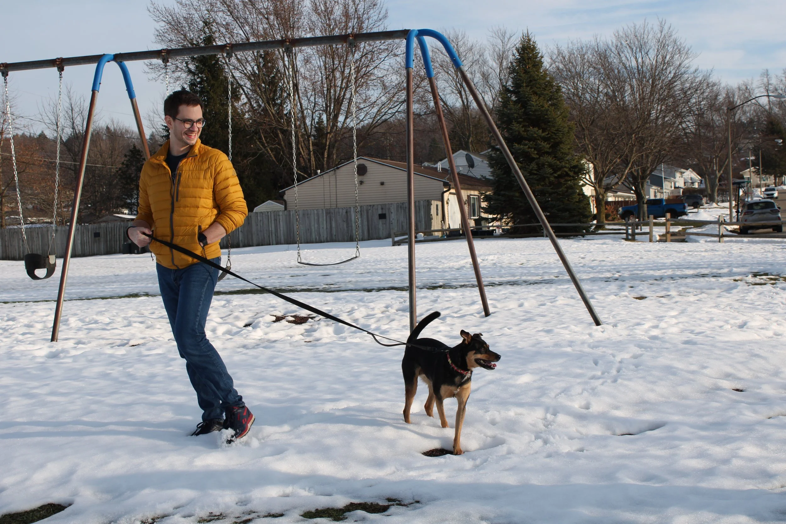 A man in a yellow jacket walking a dog on a leash in a snow-covered park with swings and residential houses in the background.