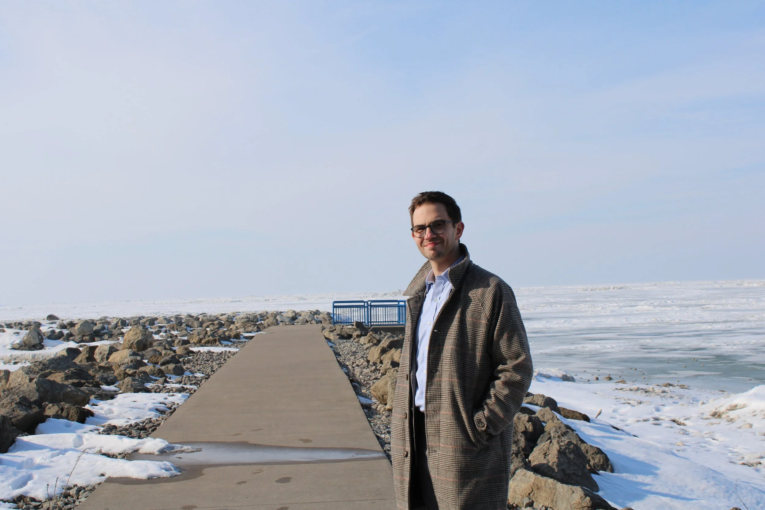 A man in a plaid coat and glasses standing on a pier by the icy lake in winter.