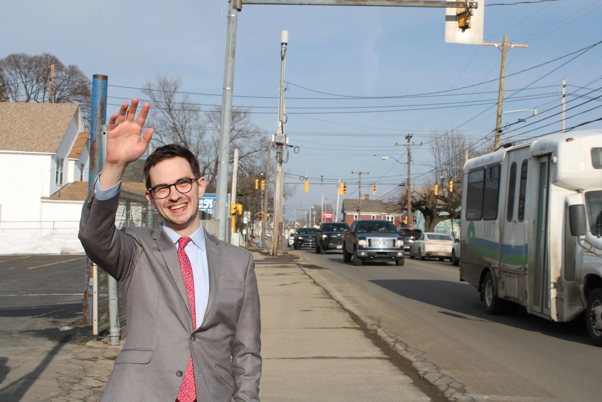 A man wearing glasses and a gray suit smiling and waving on a sidewalk in a city street with cars, trucks, utility poles, and traffic lights.