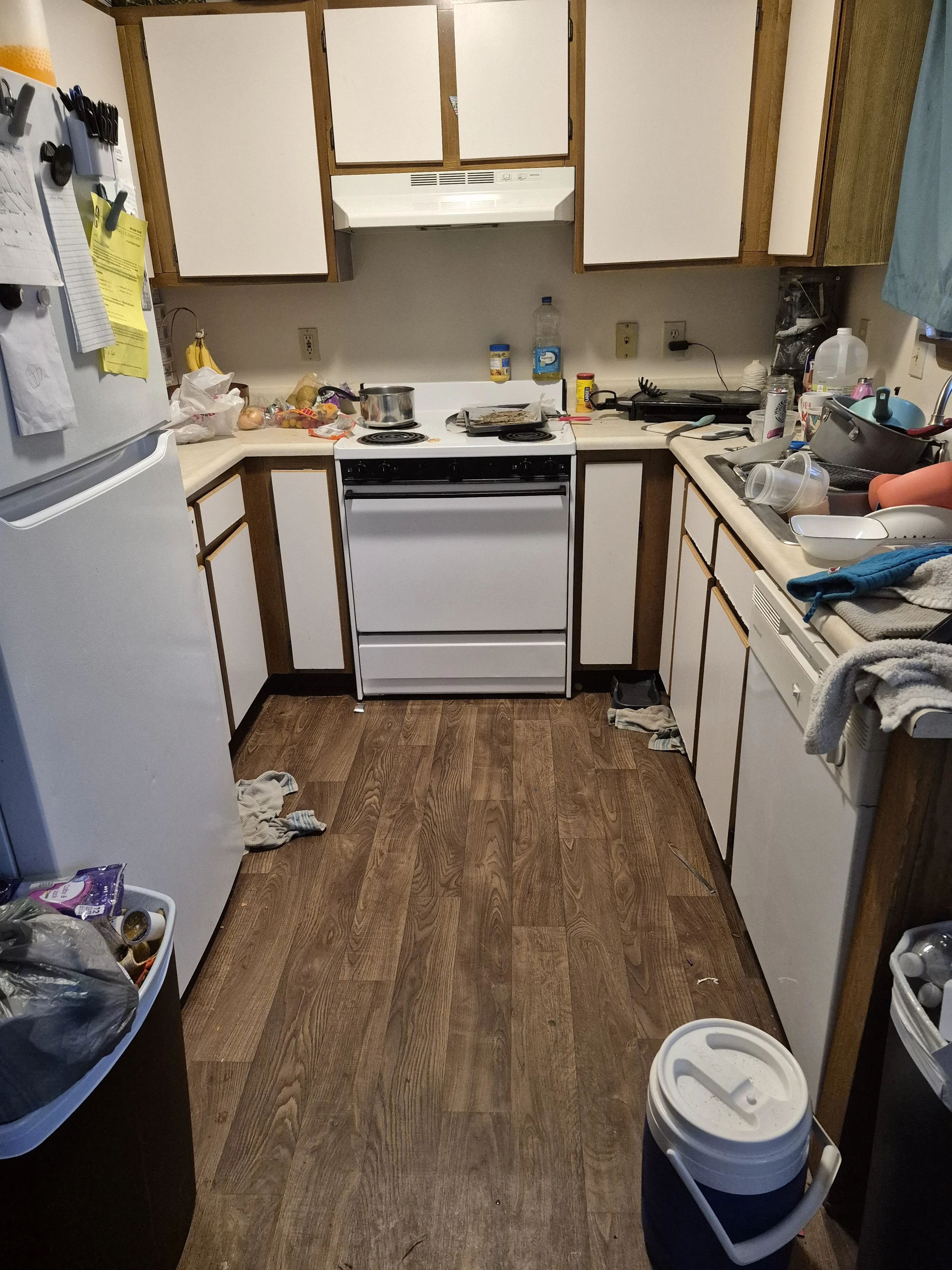 A cluttered kitchen with white cabinets, a white stove, and a refrigerator. The countertops are filled with various dishes, containers, and food items. There is a dish drying rack with dishes on the right side, and the floor has some clothes and debris.