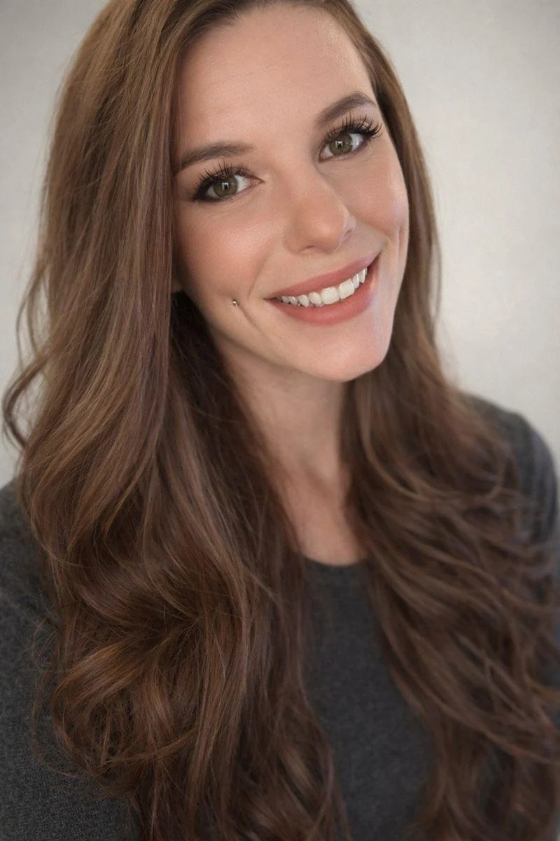 A woman with long, wavy, reddish-brown hair and green eyes, smiling and wearing a dark gray top, against a plain light background.