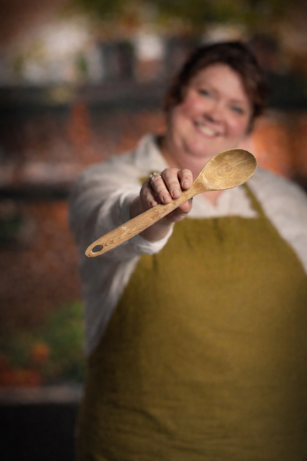 A smiling woman holding a wooden spoon towards the camera in a kitchen or cooking space.