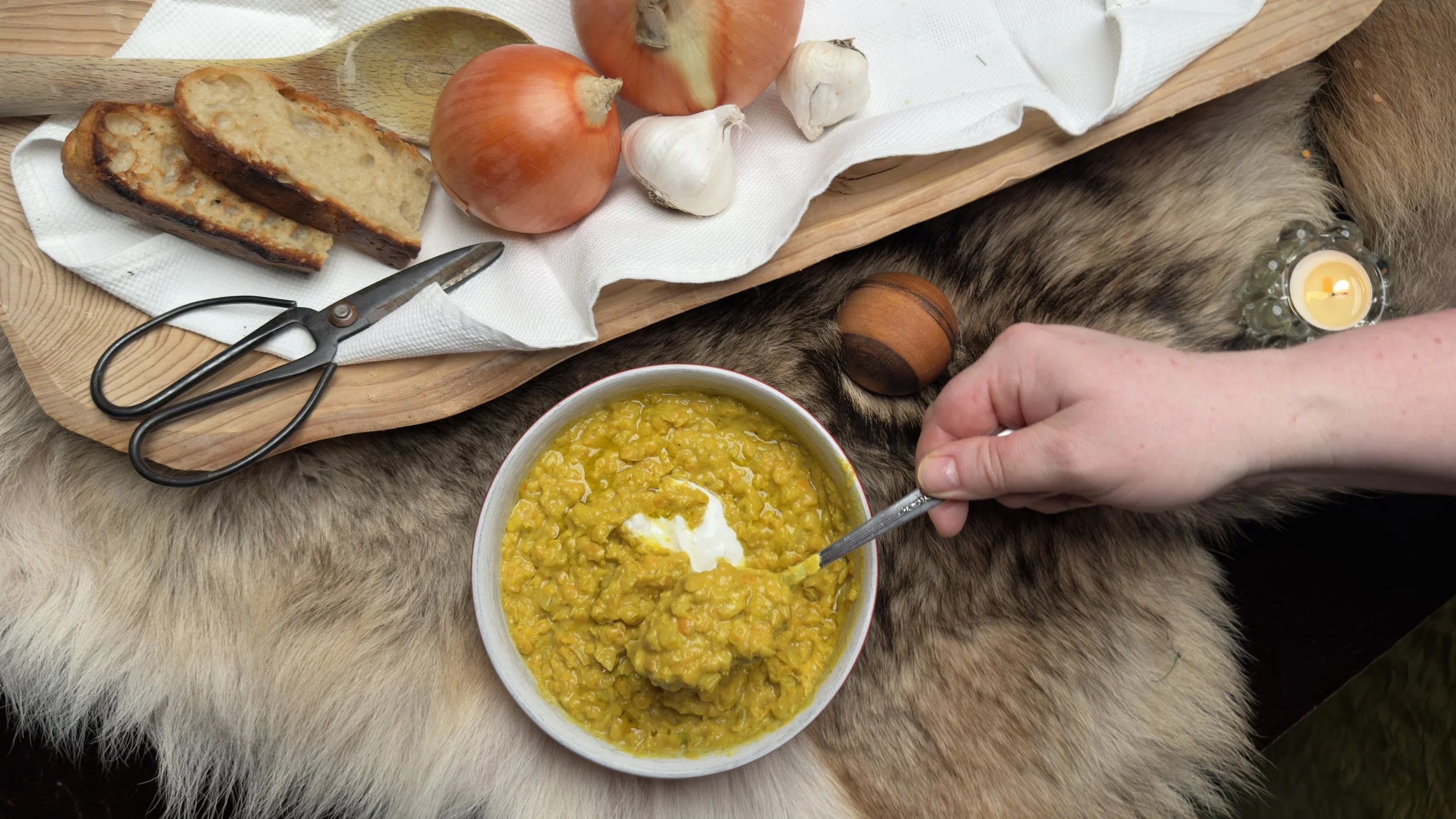 A bowl yellow lentil soup dollop of sour yogurt on top. Surrounding it are onions, garlic, a wooden cutting board with sliced bread, a spoon, and a small candle on fur. Soup grand rapids michigan