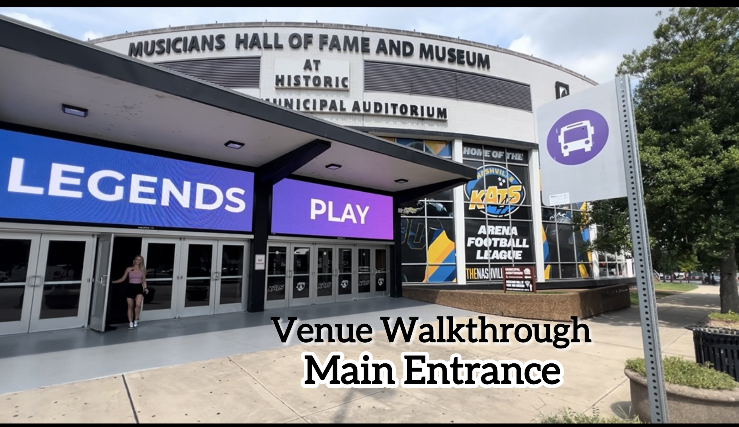 Exterior of the Nashville Municipal Auditorium with a large digital sign displaying 'Legends Play'. A person is walking out of the entrance doors, and there is a bus stop sign on the sidewalk. A banner advertising the Nashville SC football league is visible on the building.