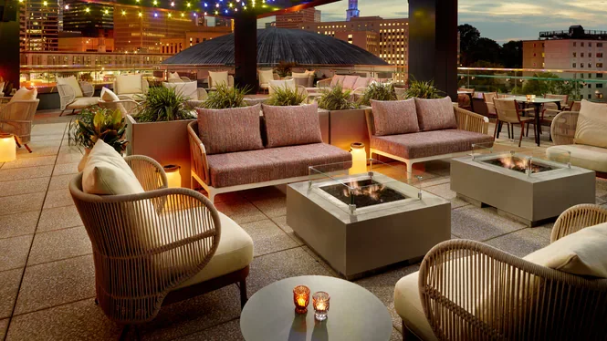 Rooftop lounge with outdoor seating, fire pits, string lights, and city skyline during evening