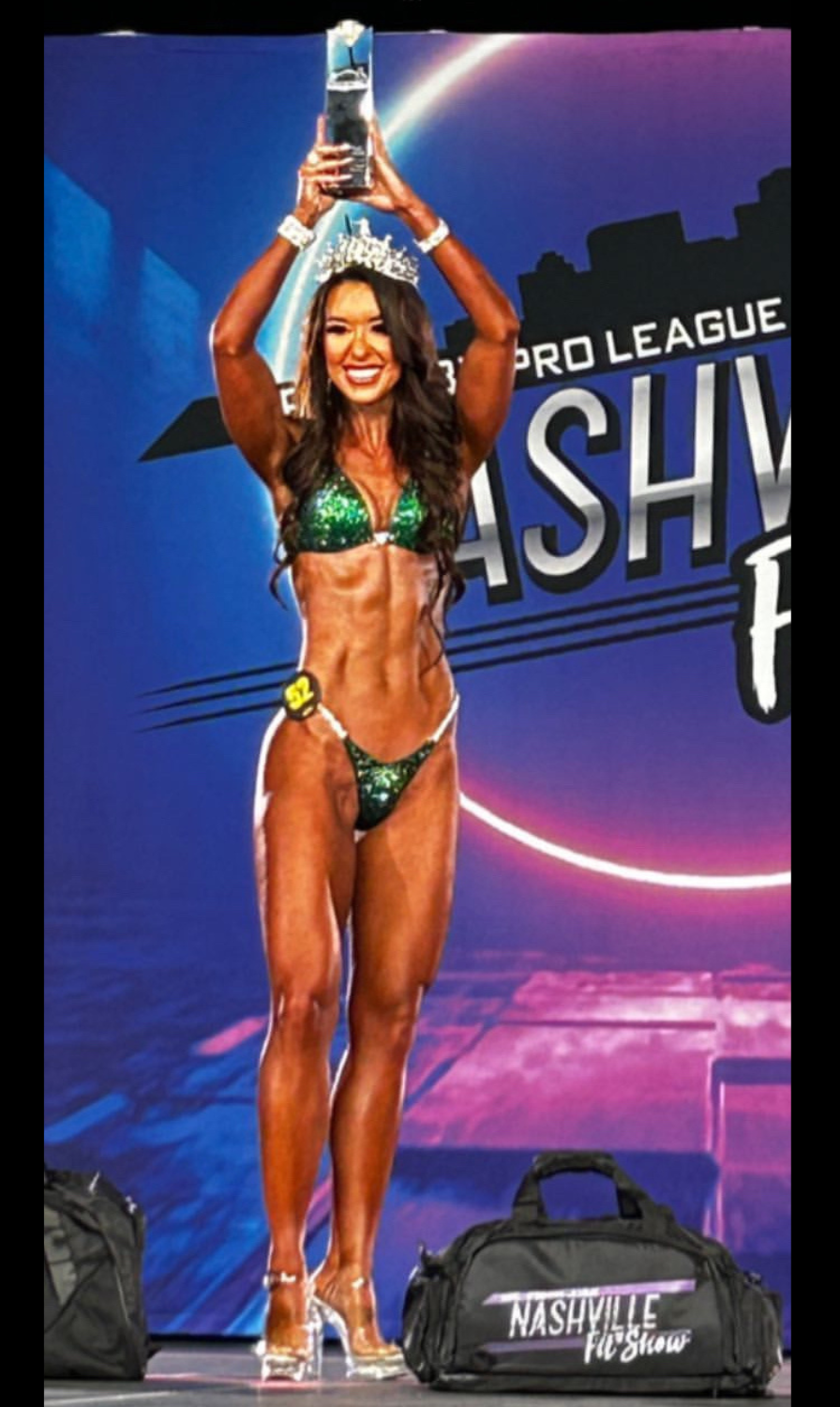 Female bodybuilder in green bikini with a crown holding a trophy on stage at a fitness competition.