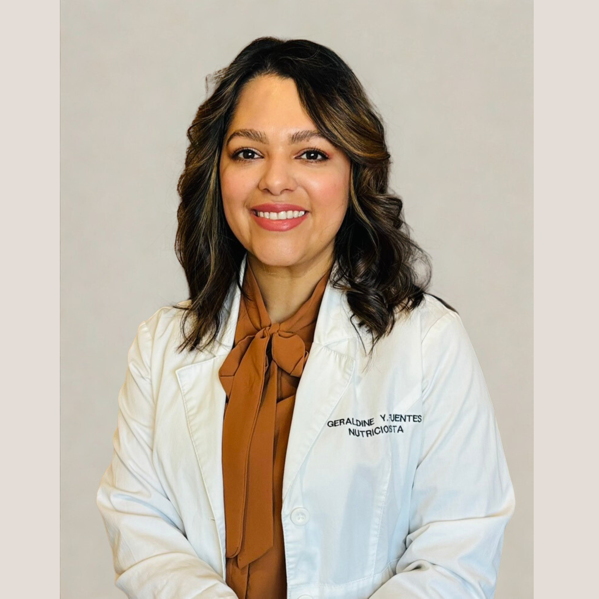 Mujer con cabello castaño y ondulado, vestida con bata blanca y una bufanda marrón, sonriendo, en un fondo gris, con la inscripción 'Geraldine Y. Juentes Nutricionista' en la bata.