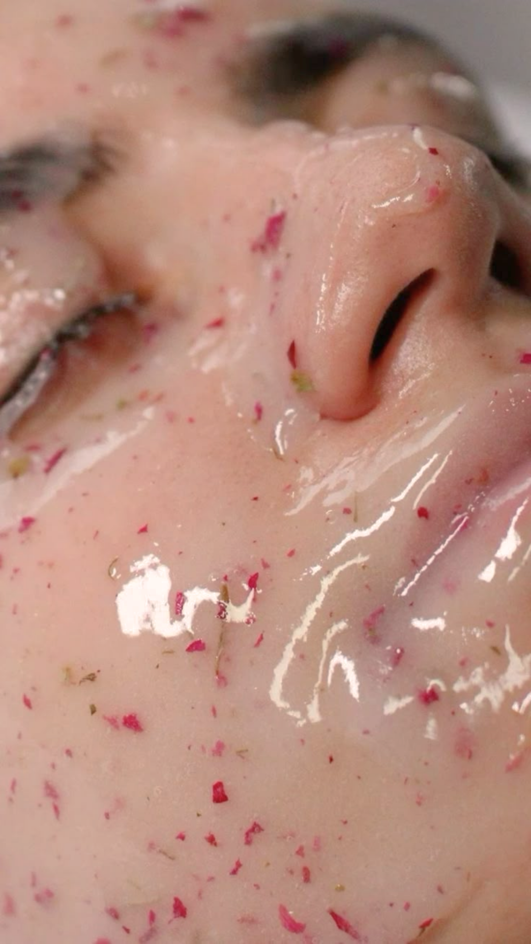Close-up of a person's face with eyes closed, covered in pink and red flower petals and some green bits, with a shiny, wet appearance.