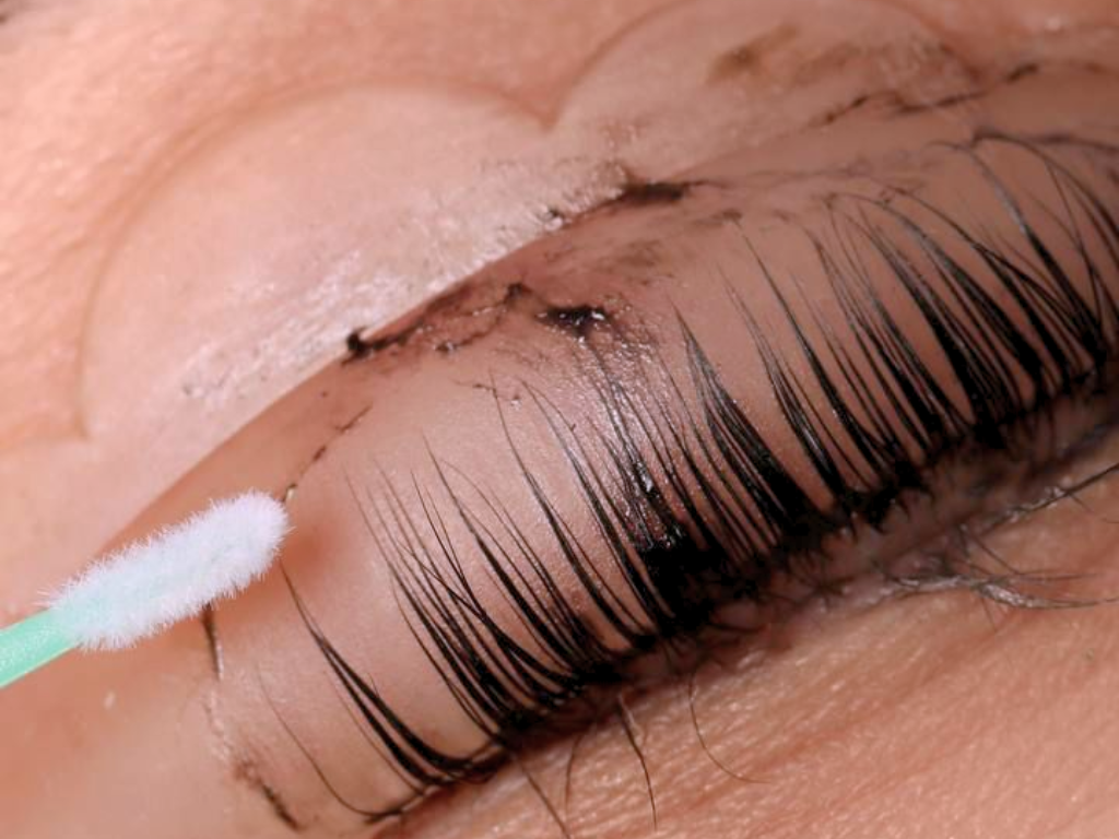 Close-up of a person's eye with long, dark eyelashes and eyeshadow. A cotton swab is being used near the eyelid, possibly for makeup application or removal.