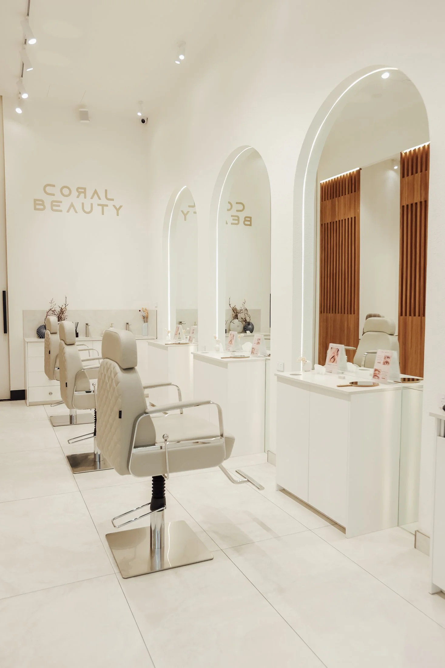 An interior of a modern beauty salon with white walls, beige salon chairs, and large mirrors. The salon has a clean, minimalist decor with wooden accents and soft lighting.