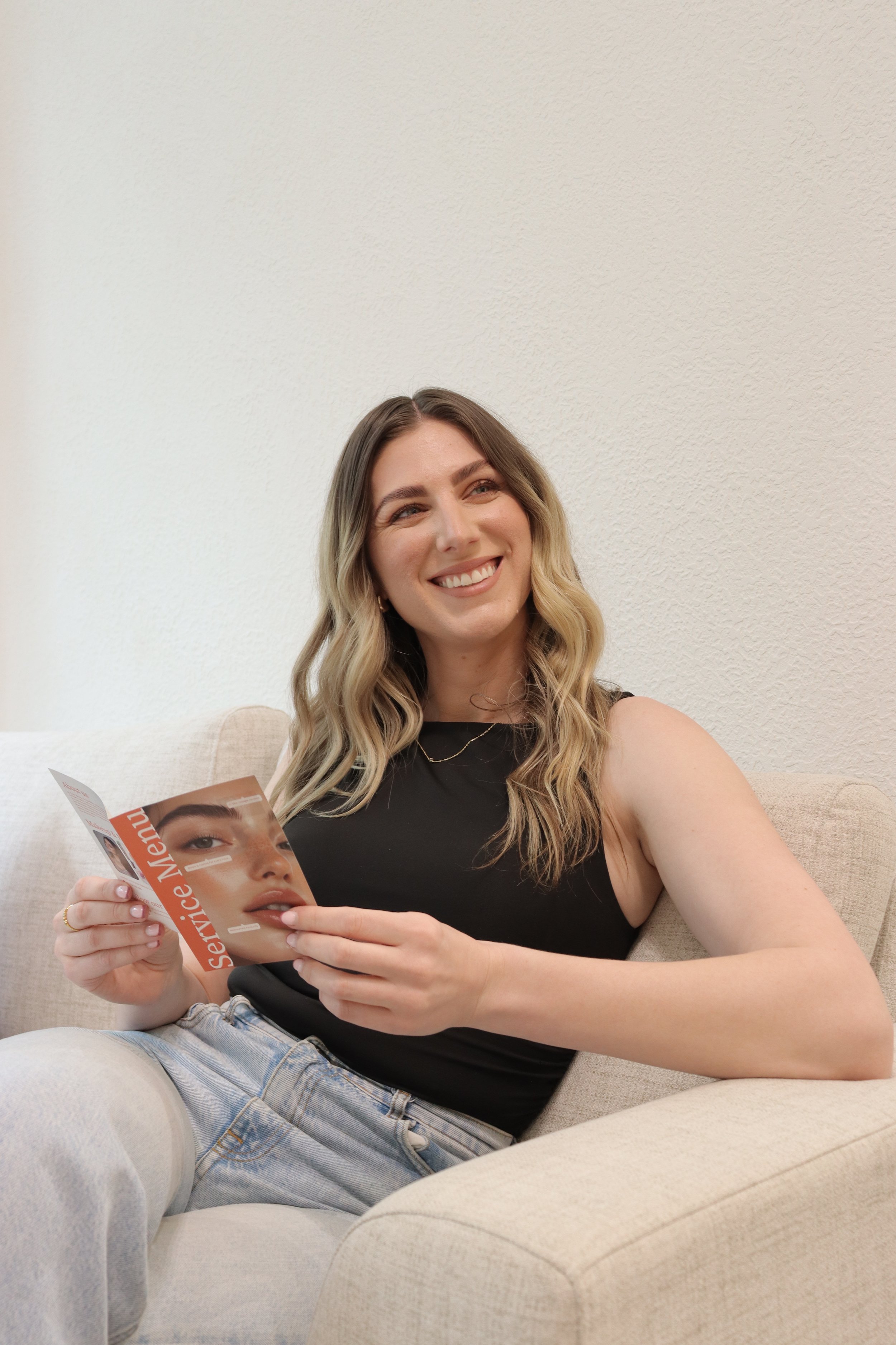 Woman sitting on a beige couch, smiling, holding a magazine with a woman's face on the cover, against a plain wall.