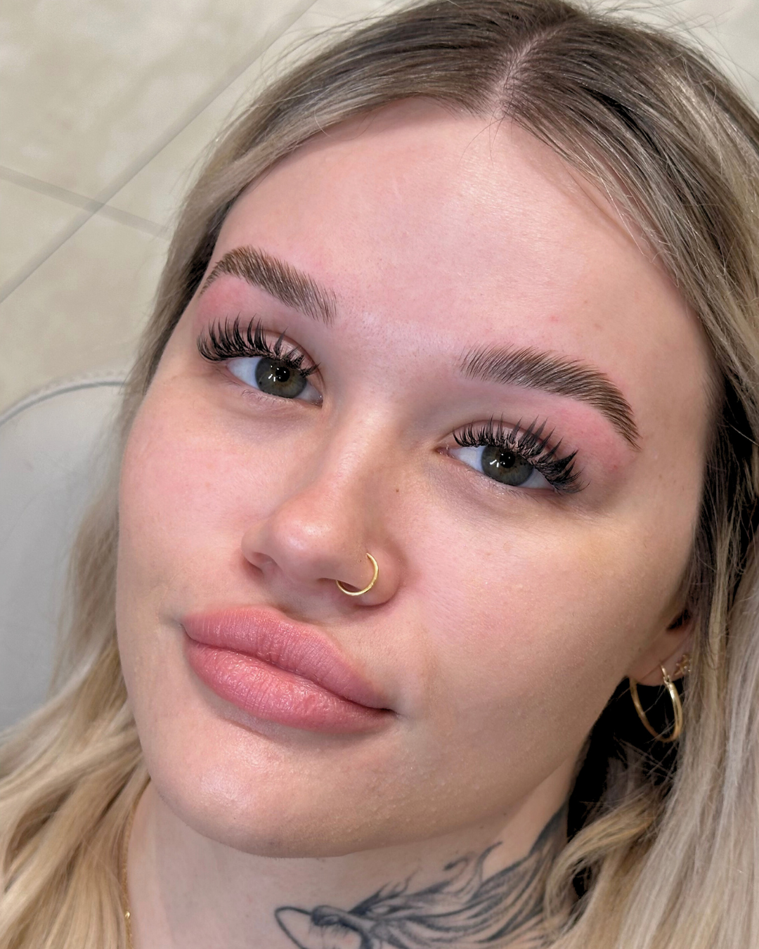Close-up of a young woman with blonde hair, green eyes, and full lips. She has well-groomed eyebrows, long eyelashes, a gold nose ring, and gold hoop earrings. There is a tattoo on her neck.