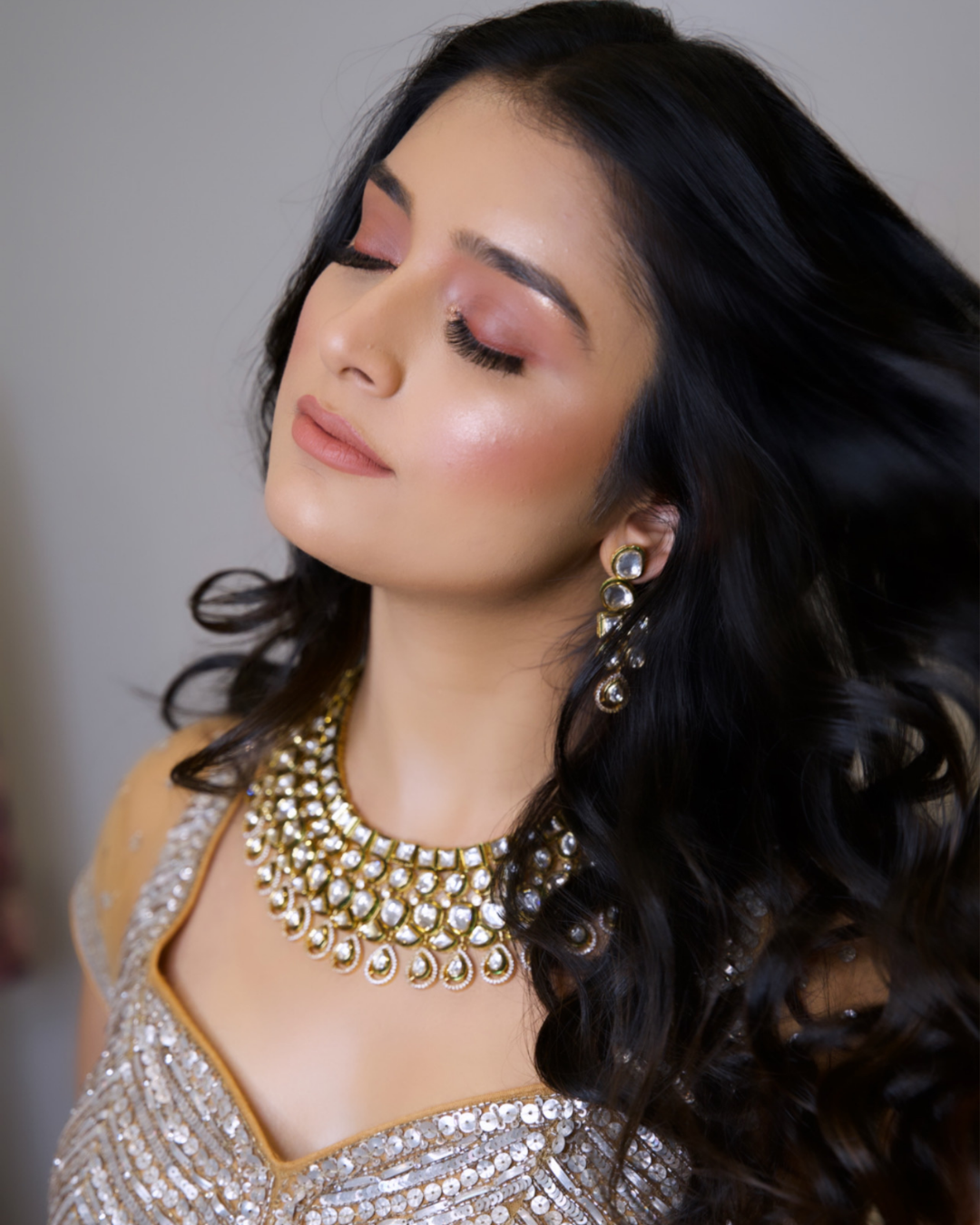 A woman with long black hair and closed eyes, wearing gold and white jewelry and a gold, sequined dress.