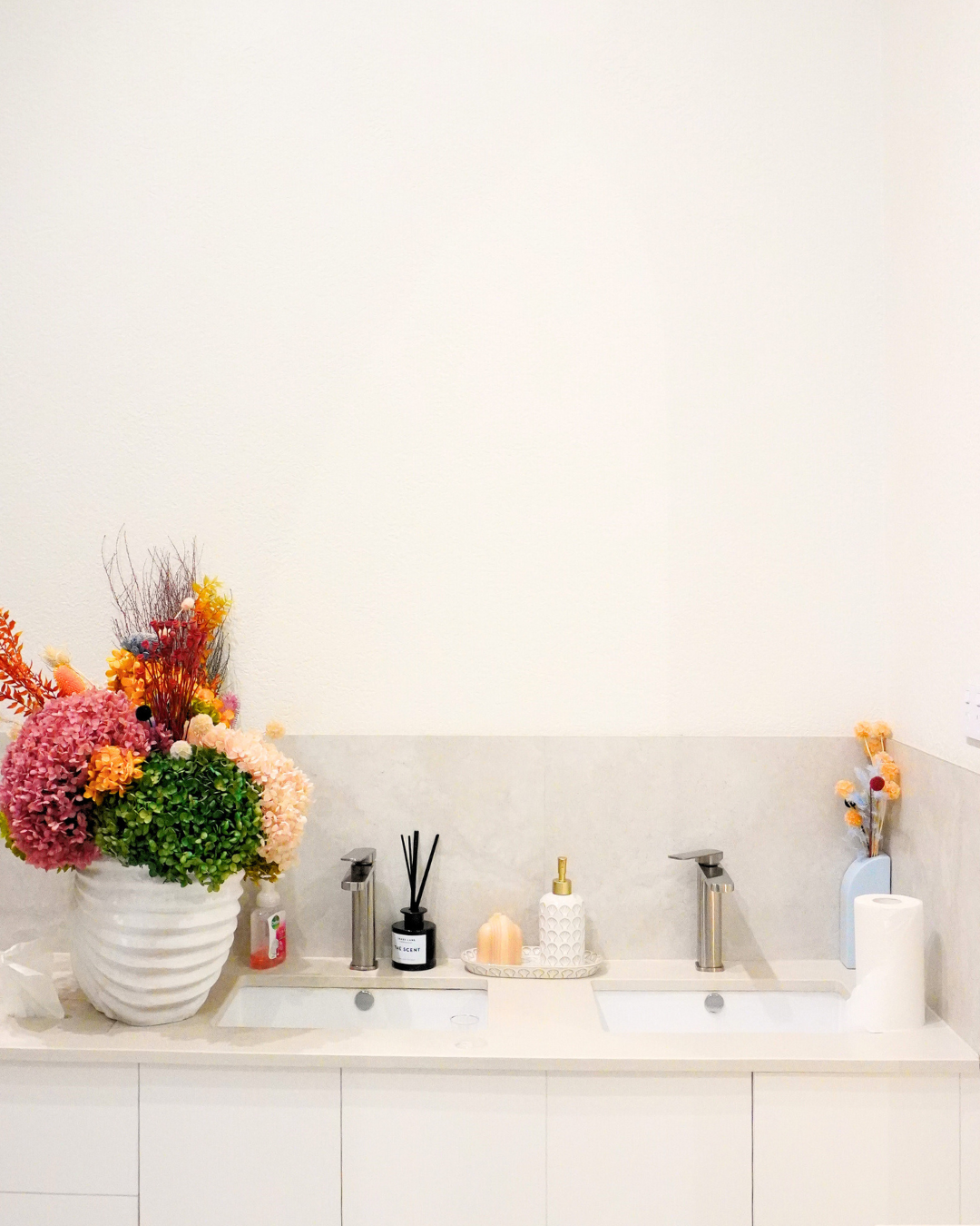 Bathroom vanity with two sinks, large flower arrangement, soap dispenser, reed diffuser, decorative candles, and paper towel roll.