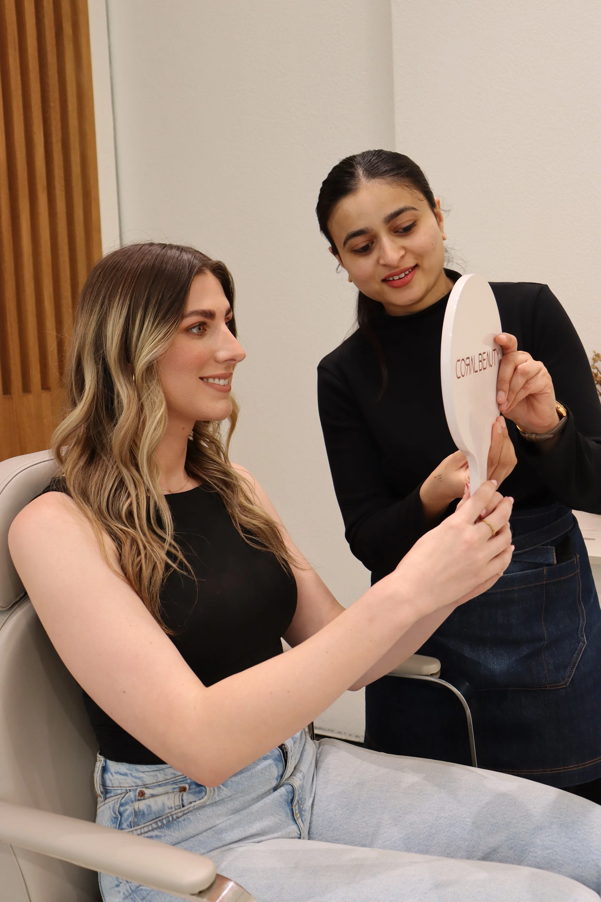 Two women, one seated and one standing, looking at a mirror together. The seated woman has wavy blonde hair and is wearing a black sleeveless top and light blue jeans. The standing woman has dark hair tied back, is in a black long-sleeved top and dar