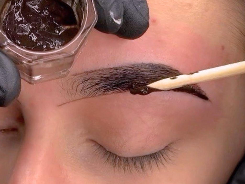 Close-up of a person getting semi-permanent eyebrow tinting with a tiny brush and dark dye.