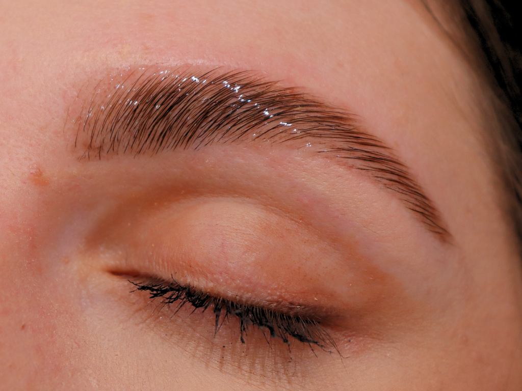 Close-up of a person's closed eye with groomed eyebrow and long eyelashes, showing smooth skin and a tiny mole near the eyebrow.