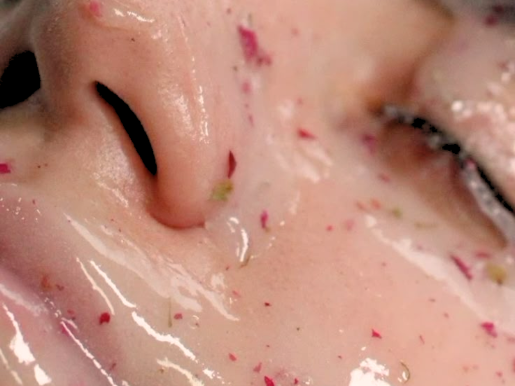 Close-up of a person's nose, mouth, and closed eye, with visible facial skin, some small red and green particles, and a shiny, moist appearance.