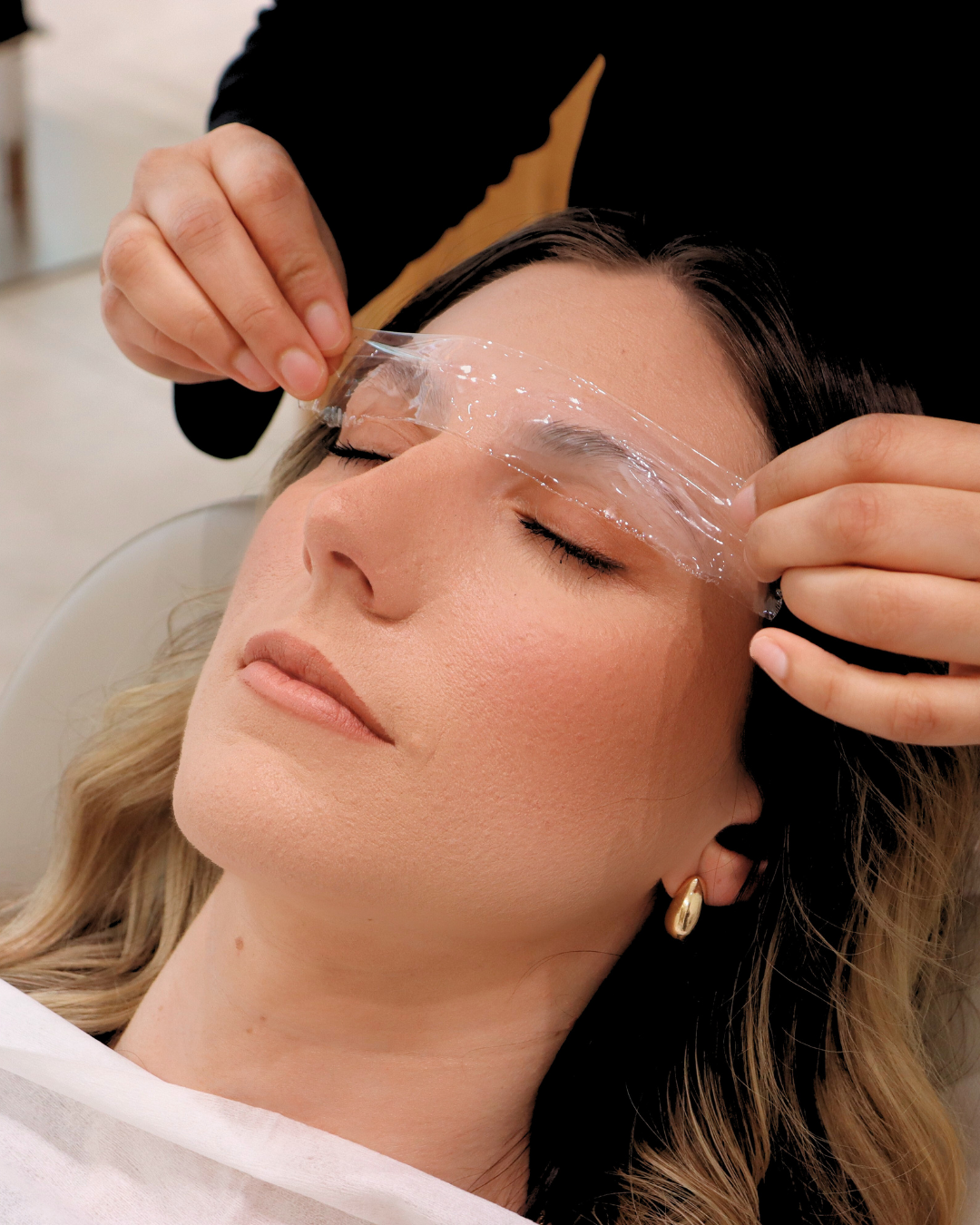 A woman receiving an eyelash lift treatment with a protective patch applied to her eyelids.