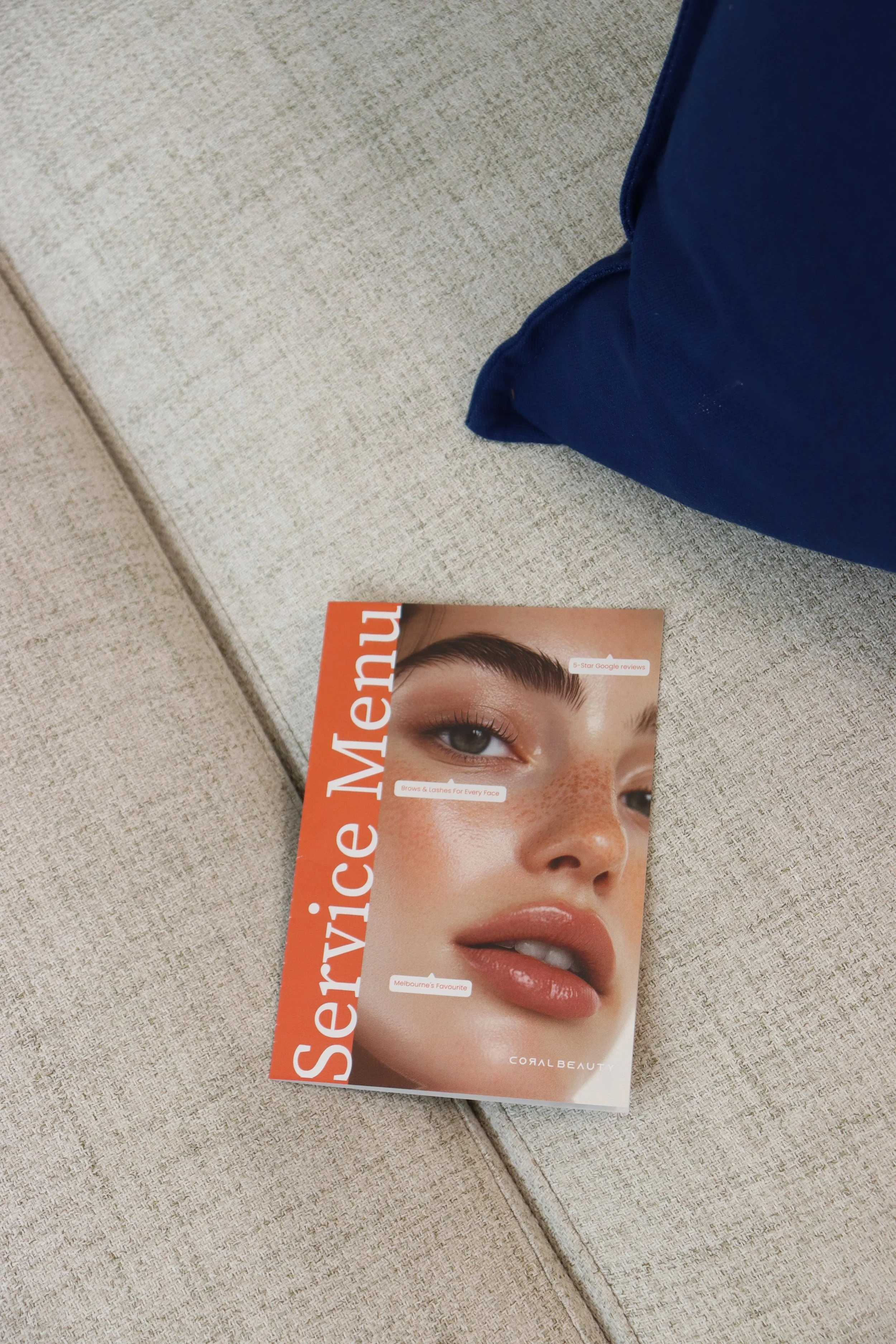 A magazine titled 'Service Menu' featuring a close-up photo of a woman's face with natural makeup, highlighting her eyebrows, freckles, and lips, resting on a beige fabric surface next to a blue pillow.