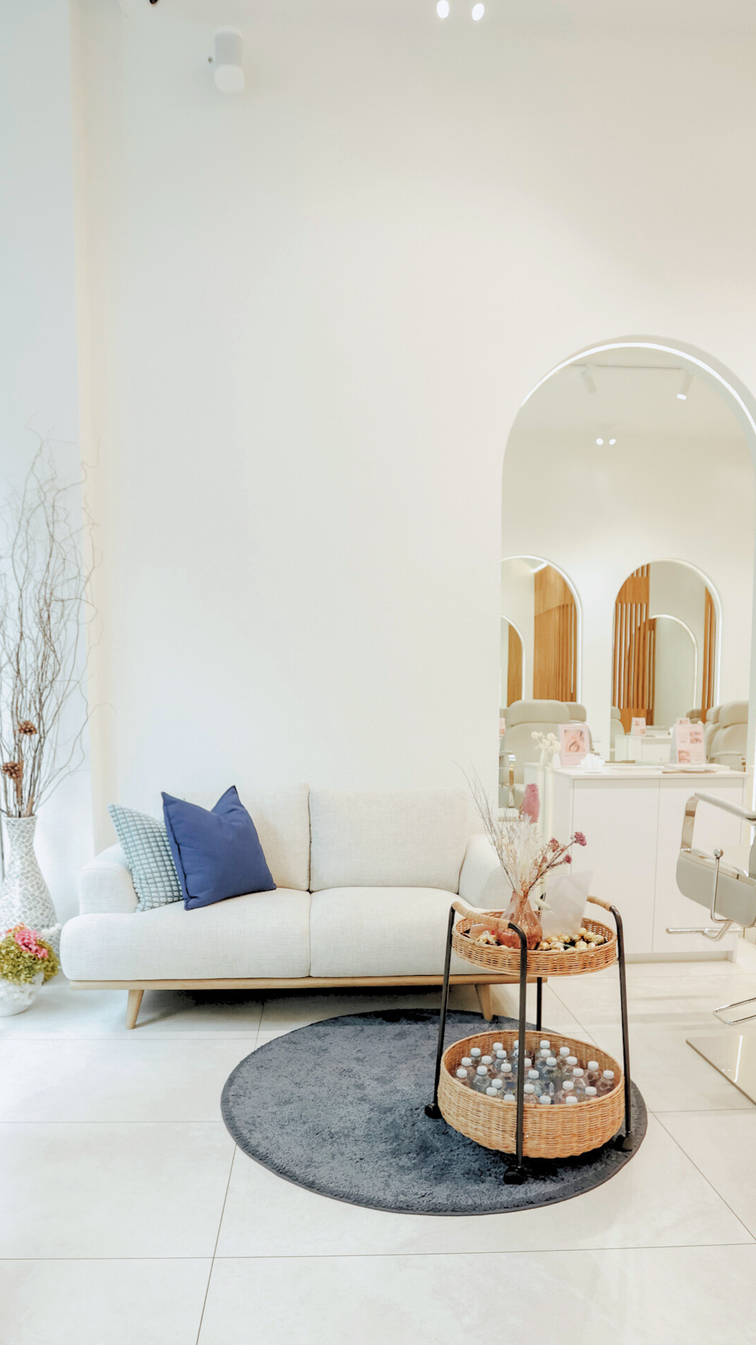 A modern, minimalistic waiting area with a white sofa decorated with blue pillows, a small round gray rug, and a two-tier wicker trolley with decorative items and bottled drinks, set against a white wall with plant arrangements.