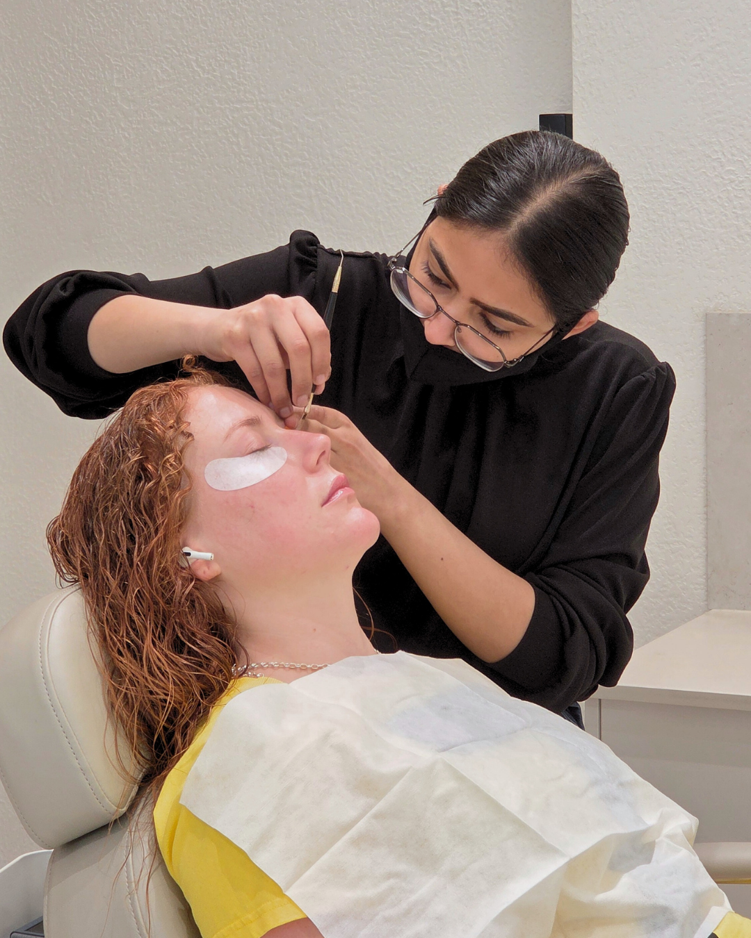 A woman with curly red hair is lying back in a medical chair with a white sheet over her chest, receiving a cosmetic procedure on her nose from a professional woman with black hair and glasses.
