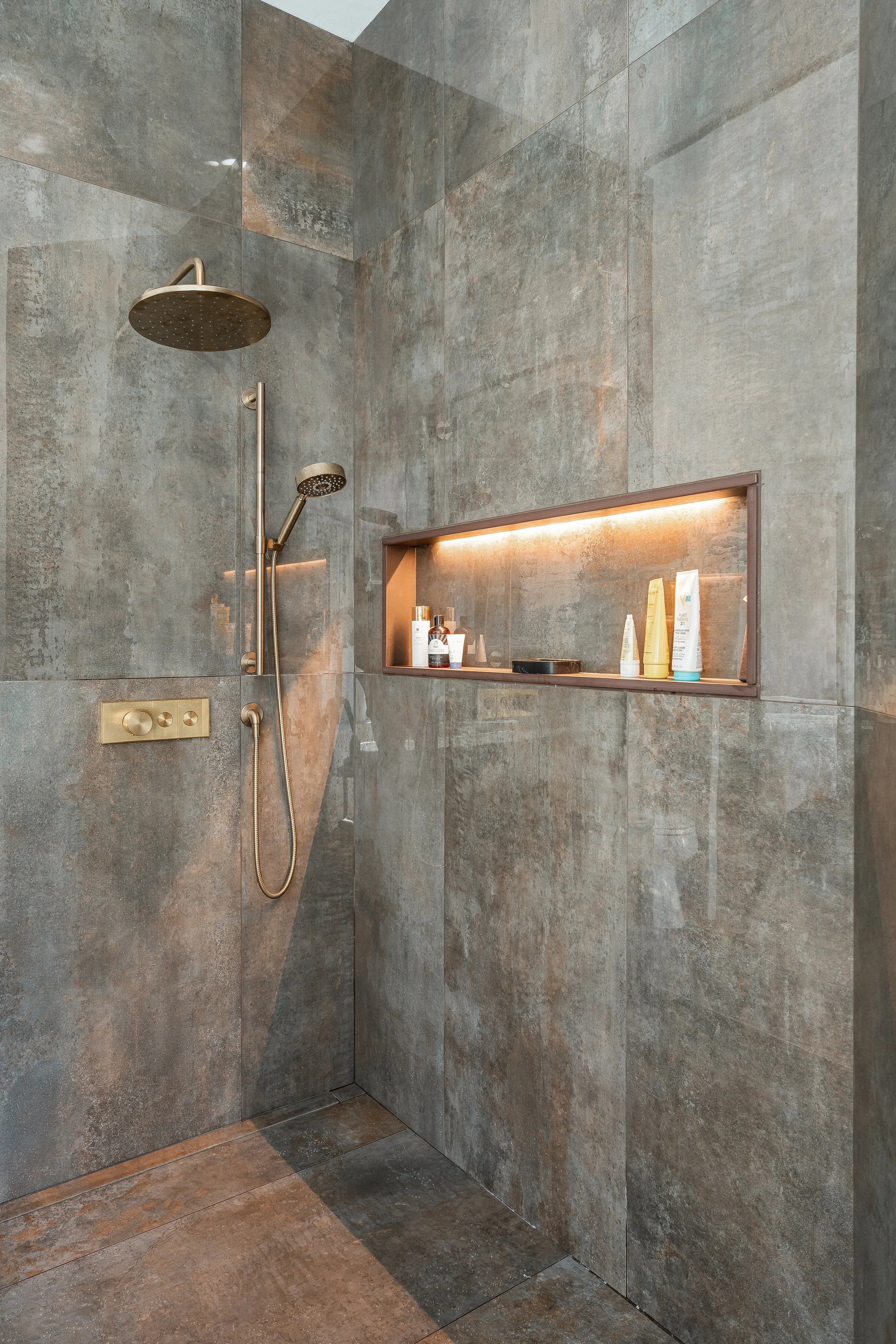 Luxury walk-in shower with gray tiles, brass showerheads, and custom niche with integrated LED lighting. 