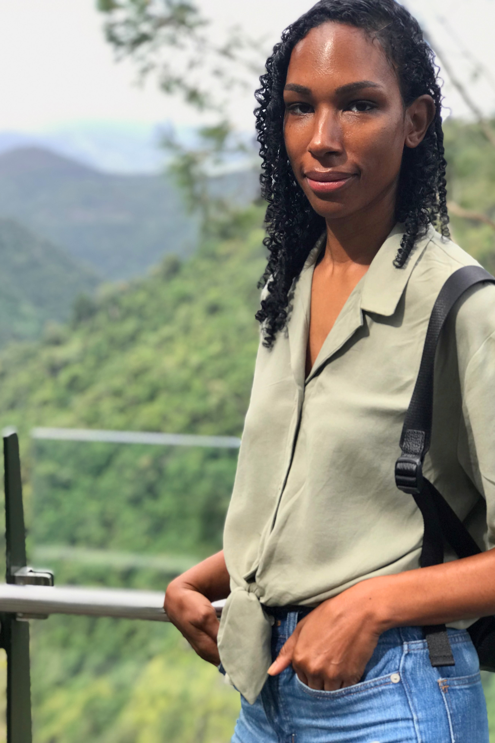 A woman with curly black hair and dark skin standing outdoors with a scenic mountainous background, wearing a light-colored shirt and jeans, with a backpack over her right shoulder.