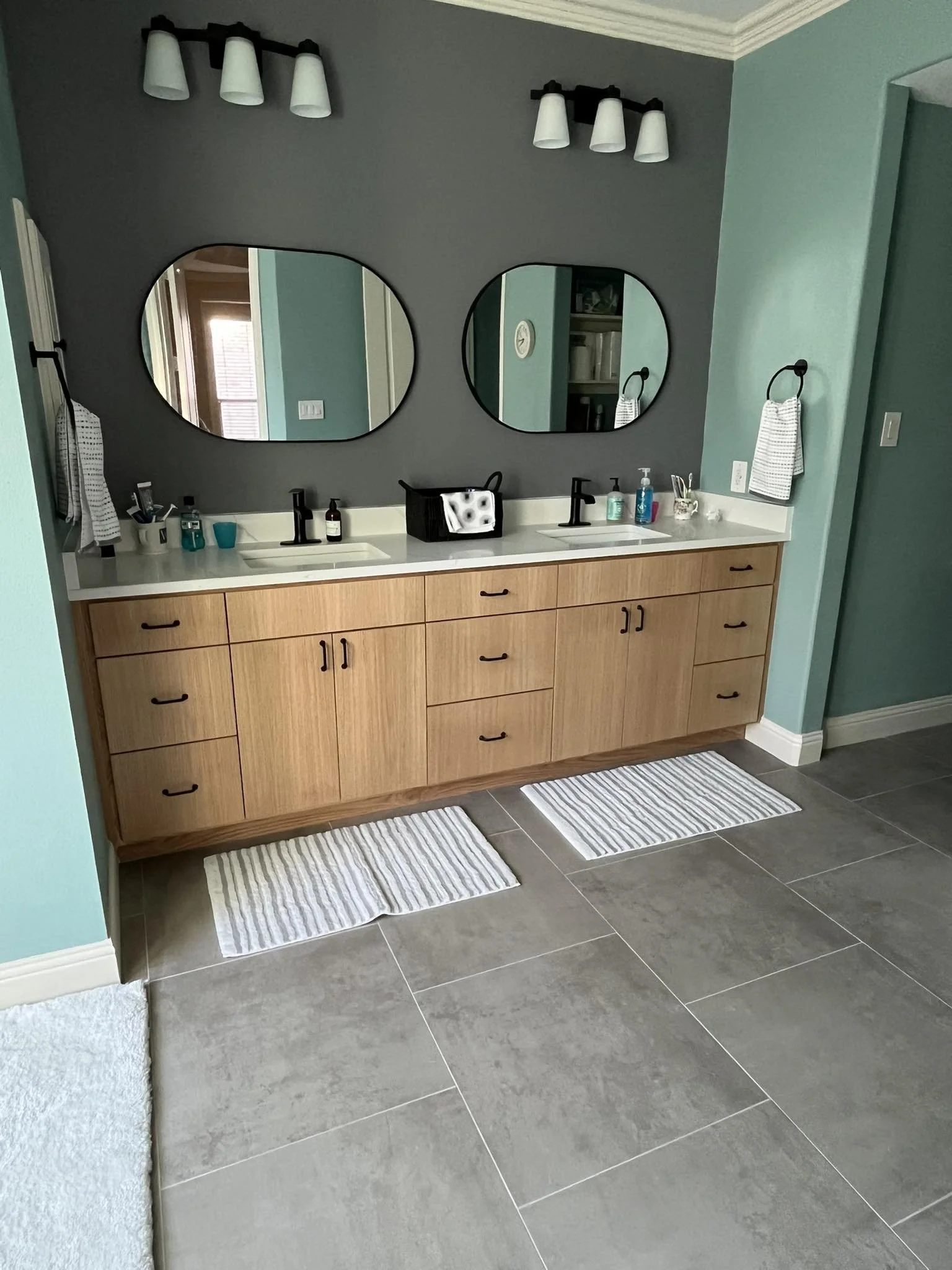 A bathroom vanity with two sinks, two oval mirrors, and wall-mounted light fixtures. The cabinet is made of light wood with black handles, and there are cleaning supplies and toiletries on the countertop. The bathroom has teal and gray walls and gray tiled flooring with two striped rugs.