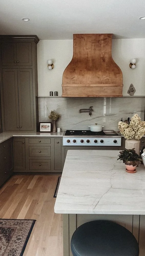 Kitchen with gray cabinets, white marble countertops, a wooden range hood, and a backsplash. Decorative plants and framed pictures are on the counter, and there are wall sconces on either side of the hood.
