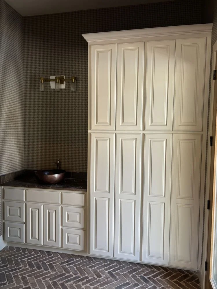A white built-in cabinet and closet with paneled doors, a dark countertop with a round bronze sink, a small backsplash, and a wall-mounted light fixture with multiple bulbs, in a room with brick-patterned flooring and dark and patterned wallpaper.