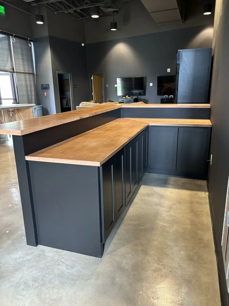 Modern kitchen with dark cabinets, wooden countertops, and an open layout leading to a living area with TV and sofa.