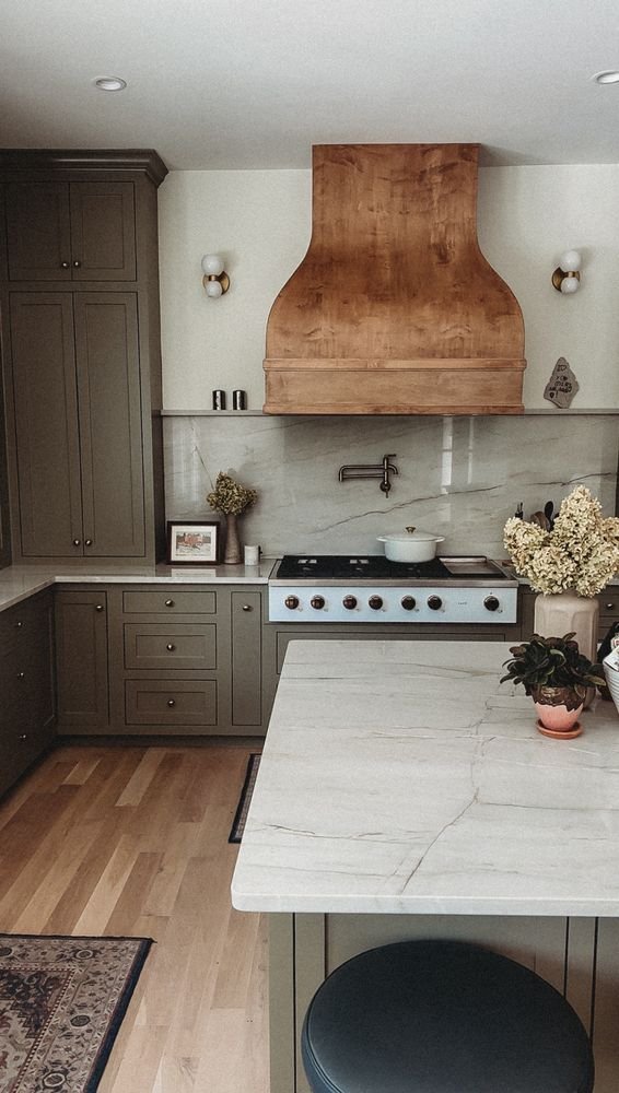 Modern kitchen with gray cabinets, a wooden range hood, white marble backsplash, and a white marble island with plants on it.
