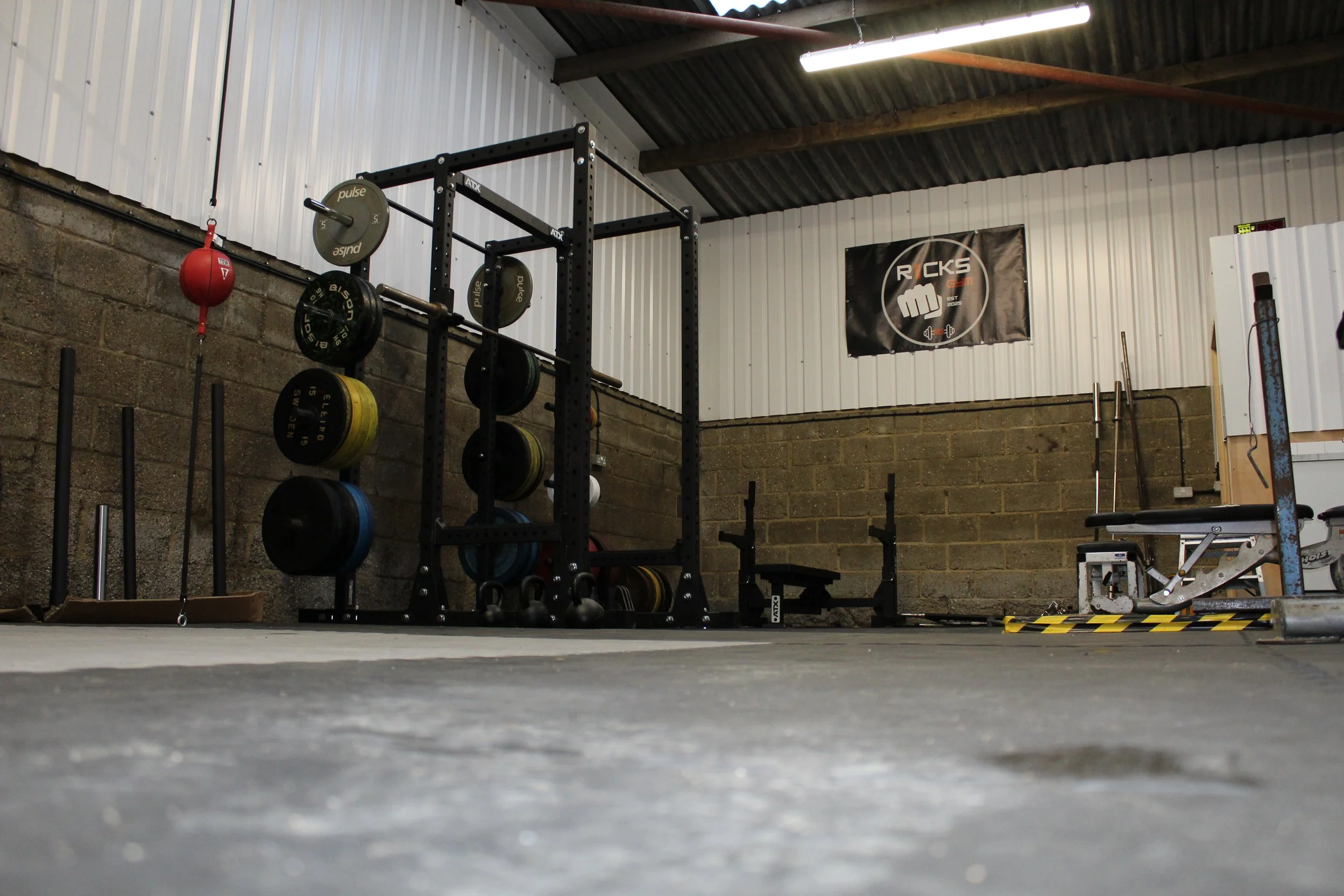 A gym space with weightlifting equipment, including a squat rack with weight plates, kettlebells, and benches. The walls are made of concrete and metal panels, and a rally-style banner with a fist logo is hanging on the wall.