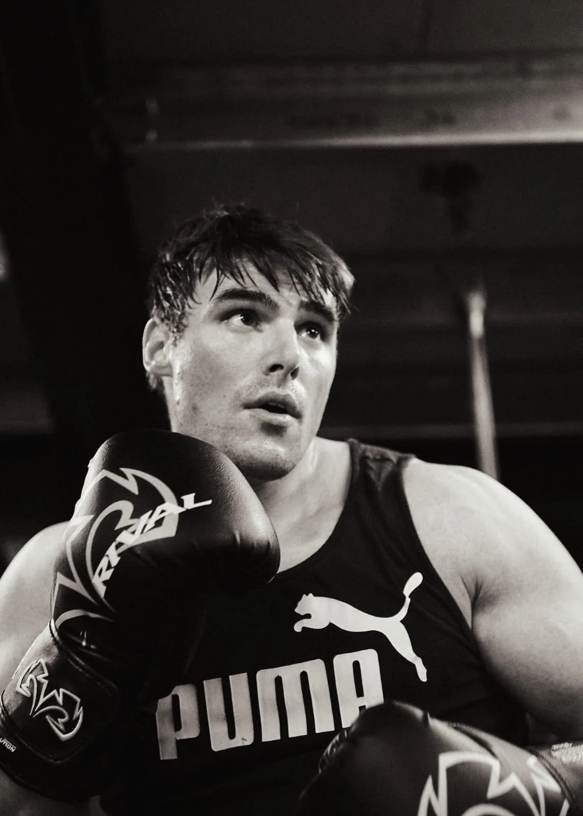 Black and white photo of a male boxer wearing gloves and a athletic top with the Puma logo, looking contemplative.