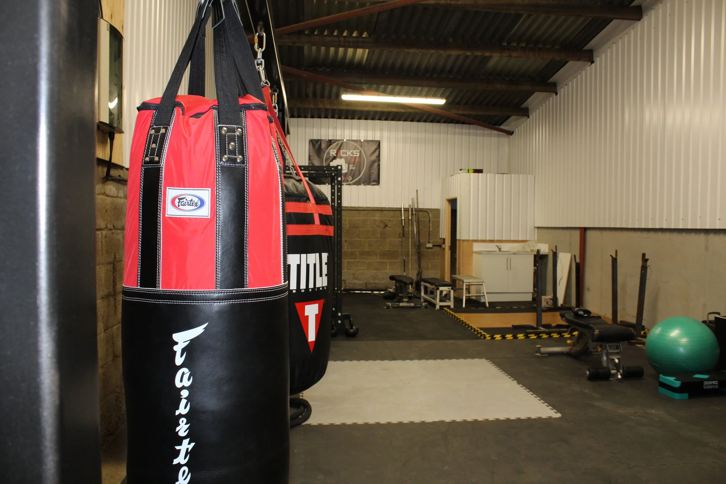 Inside a gym with boxing and fitness equipment, featuring a red and black punching bag in the foreground, workout benches, a stability ball, a medicine ball, and a small kitchen area in the background.