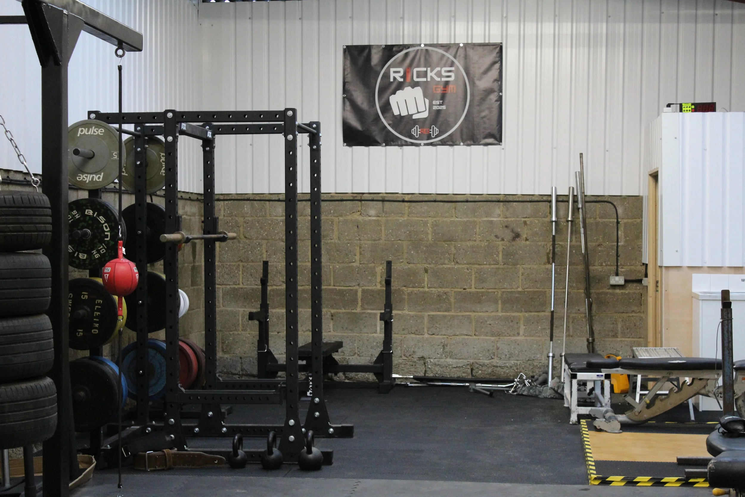 Inside a gym with black rubber flooring, a black metal squat rack with weight plates, medicine balls, and kettlebells nearby. There is a white bench with a weightlifting bar, a white wall-mounted whiteboard, and a sign with a fist logo reading "Ricks