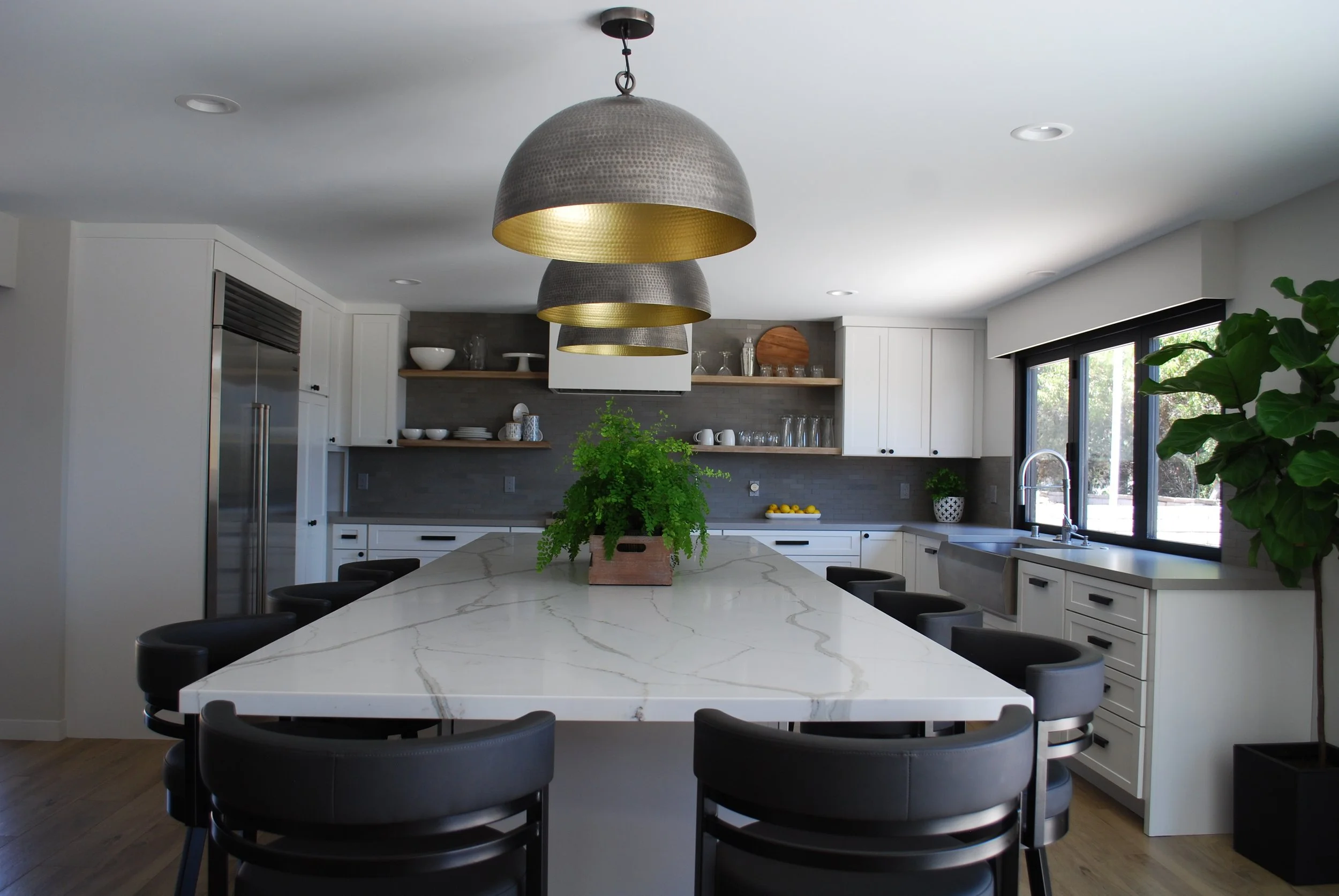 Modern kitchen with white cabinets, black bar stools, a large marble island, pendant lights, open shelving, a window overlooking greenery, a green potted plant, and a farmhouse sink.
