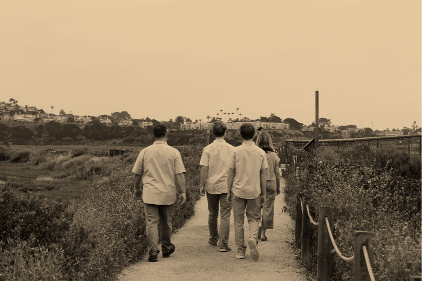 Four people walking along a trail in a park or nature area, with houses and palm trees in the background, in a sepia tone image.