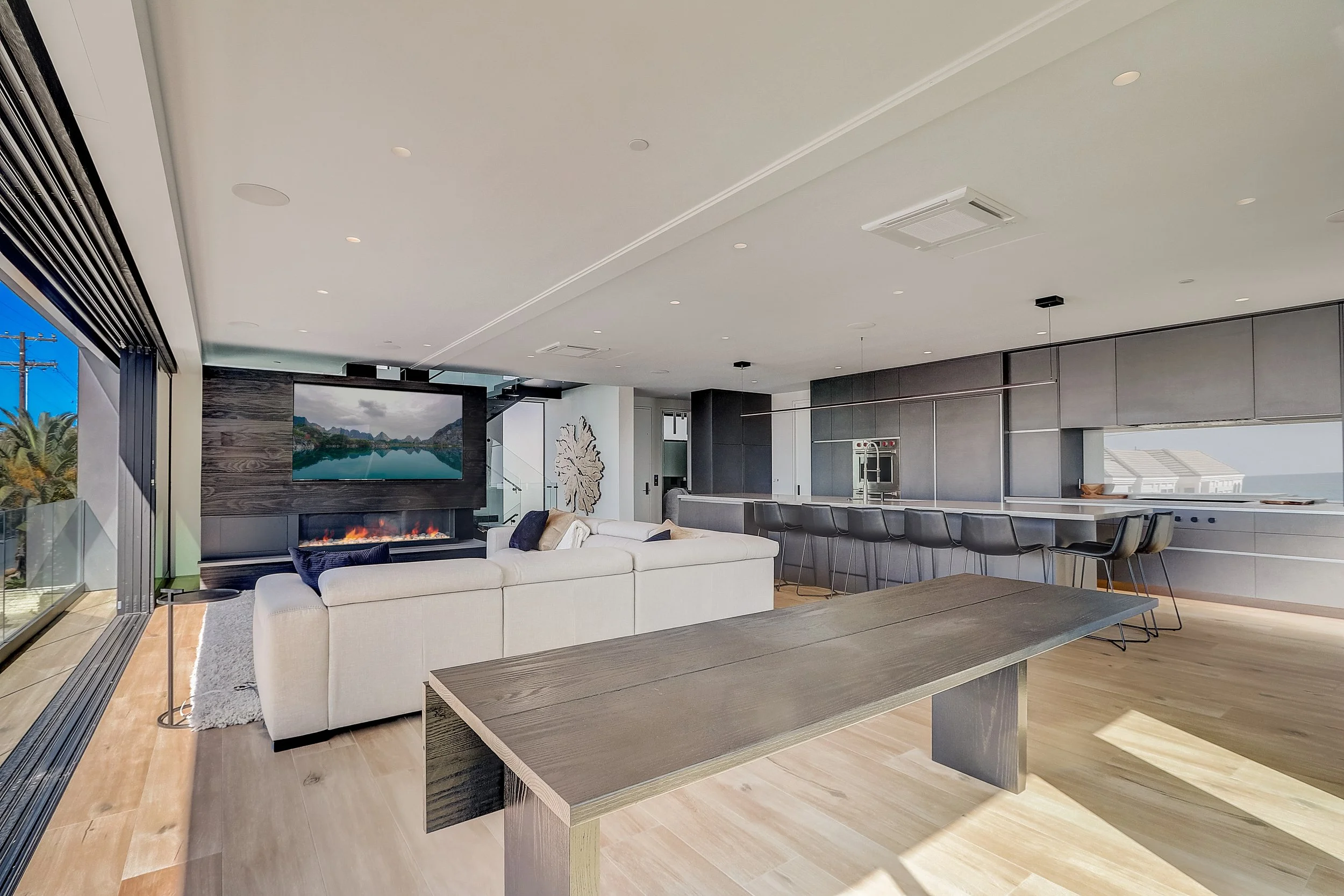 Carlsbad, CA open-concept living and kitchen design featuring a modern fireplace, expansive island seating, and seamless indoor-outdoor flow by Anne Flynn Designs.