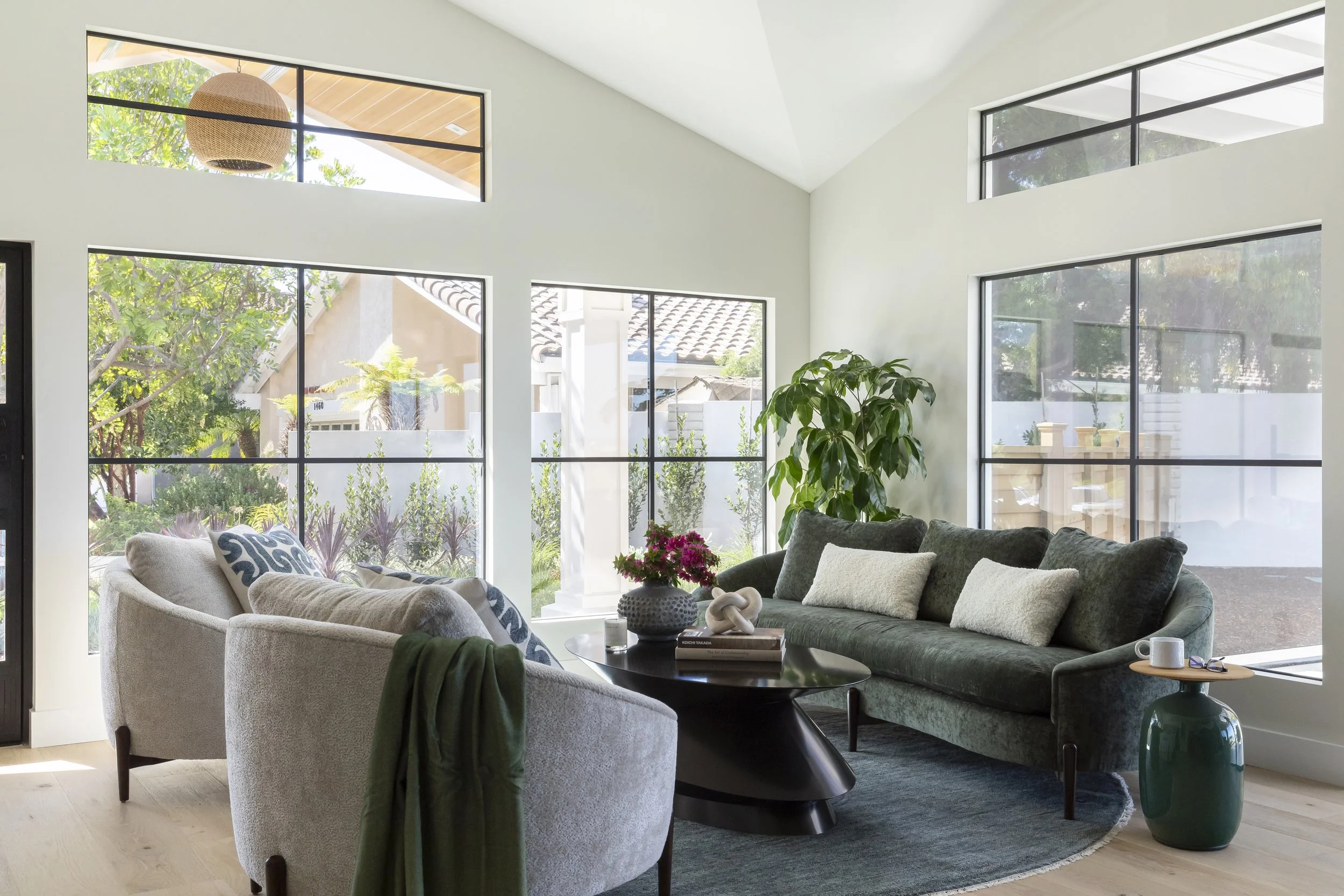 Living room with large windows, green sofa with white pillows, white armchair with blue patterned pillows, black coffee table with flowers and books, side table with mug, and green vase with plant, bright natural light.