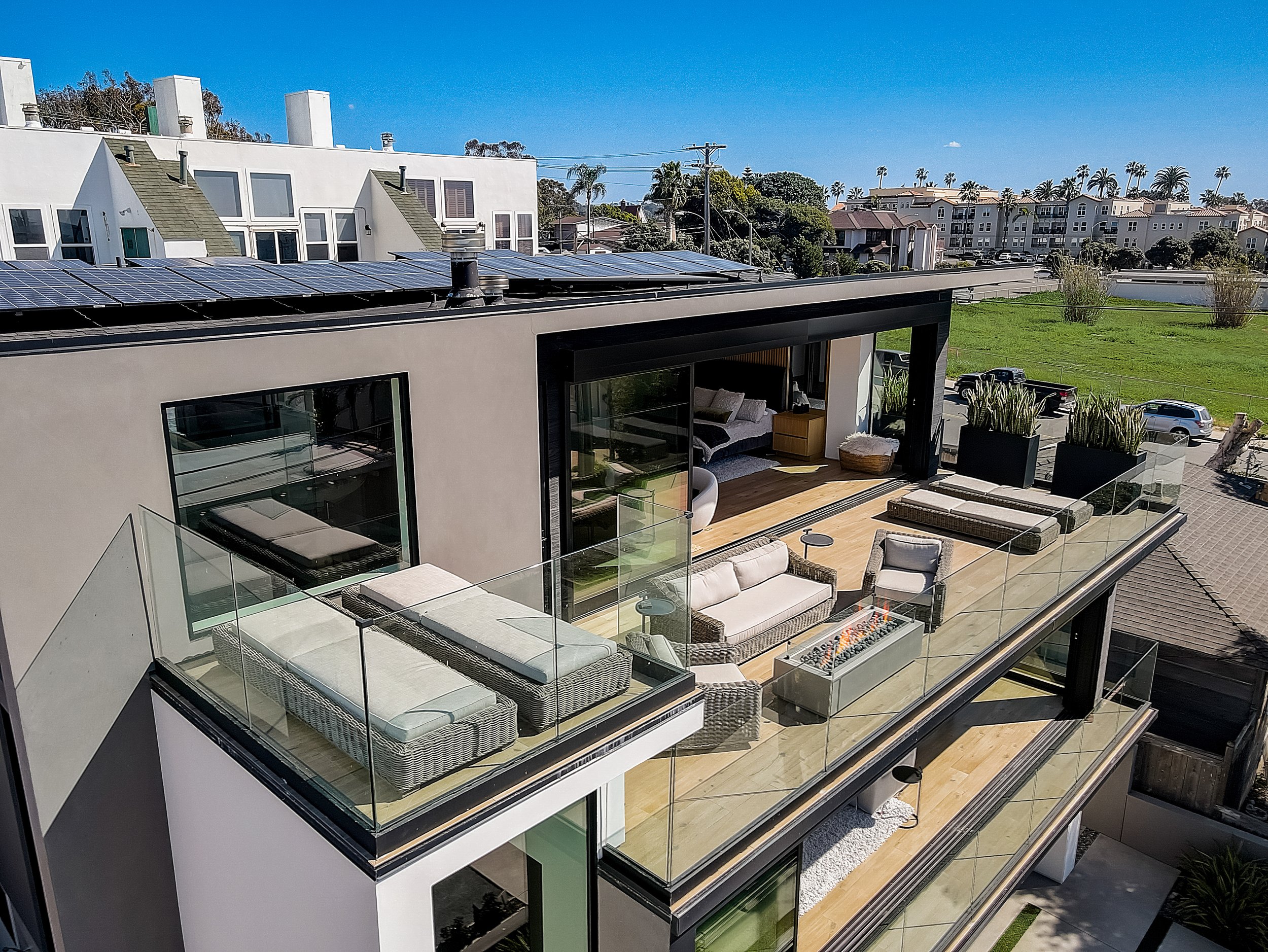 Modern rooftop terrace with outdoor furniture, glass railing, and potted plants, overlooking a suburban neighborhood with solar panels on a nearby building.