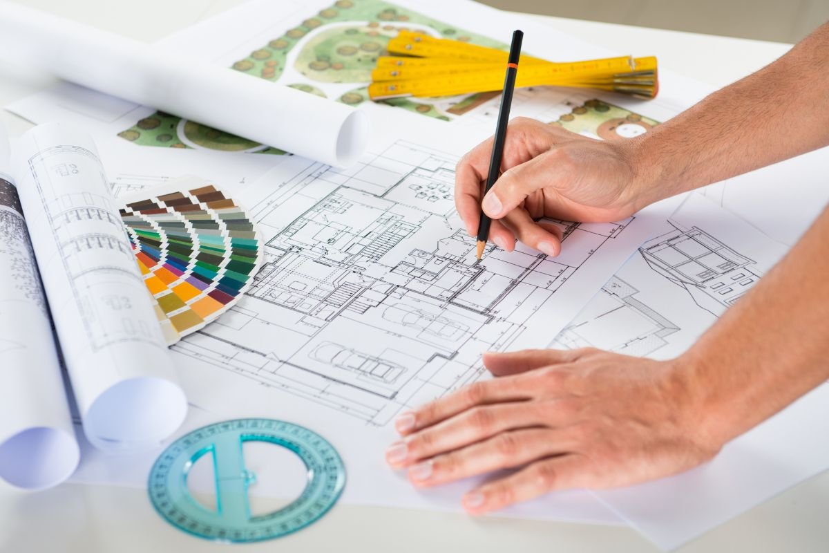 An architect's hands reviewing a building blueprint with color swatches, charts, and a yellow measuring tape on the desk.