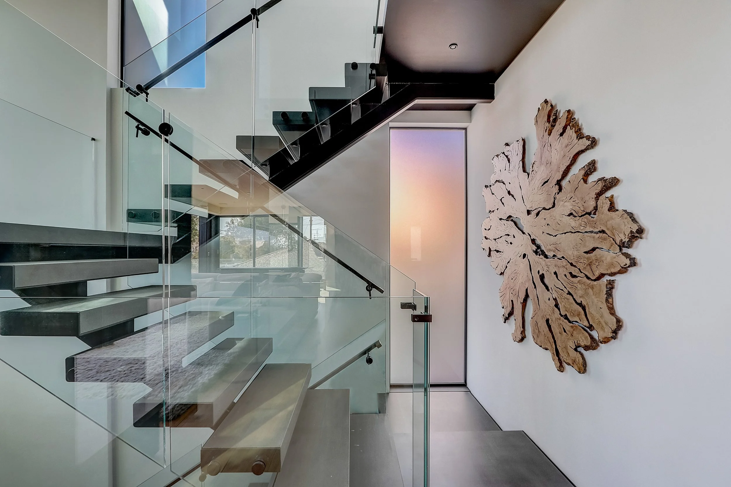 Carlsbad, CA stairwell design featuring a floating staircase with glass railings, sculptural wall art, and clean modern detailing within a contemporary beach retreat by Anne Flynn Designs.