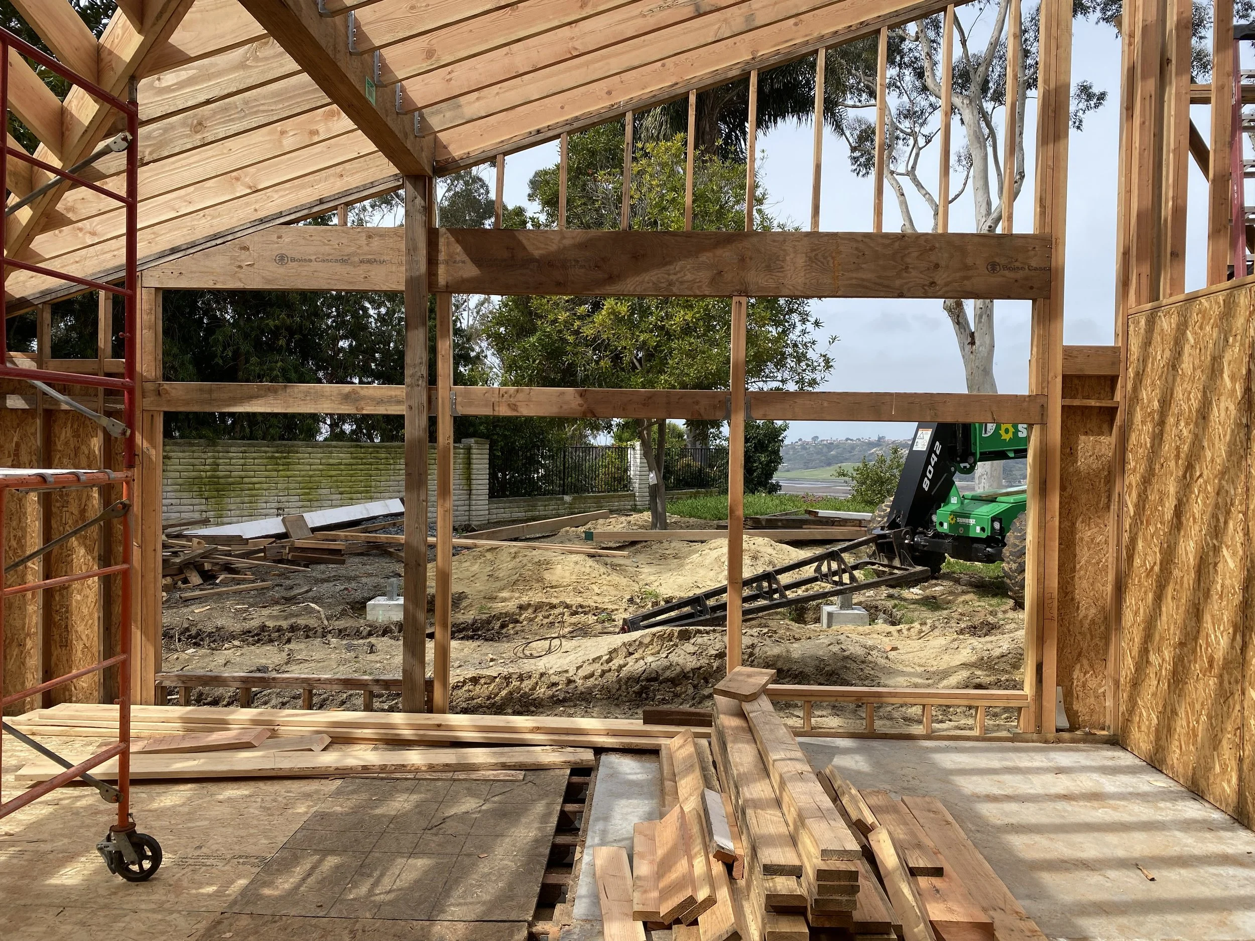 Construction site of a wooden building with exposed framing, plywood walls, scattered lumber, and a small green crane in the background outside.